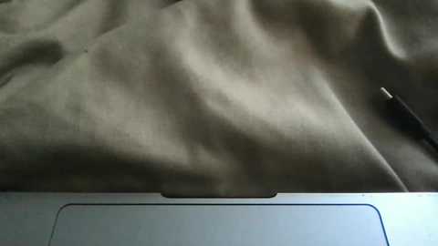 A video of a closed laptop with a black USB cable plugged into its side, resting on a wrinkled, light grey fabric surface. The image captures a minimalist, utilitarian setup.
