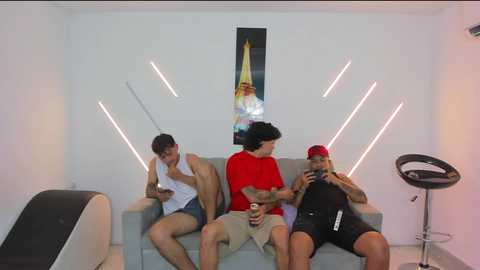 Video of three men sitting on a light grey couch in a minimalist room with modern decor. Two men in casual attire, one in a red shirt, watch a man in a black hat and tank top drink beer.