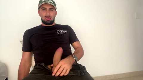 Video of a bearded, muscular, light-skinned man with a circumcised penis, wearing a black t-shirt and green cap, sitting against a plain white wall.