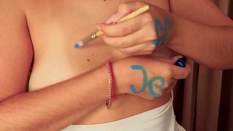 Video of a fair-skinned person applying blue body paint to their forearm, with the letters \"J\" and \"J\" visible. The person is topless, wearing a red bracelet. Background is blurred, indoors.