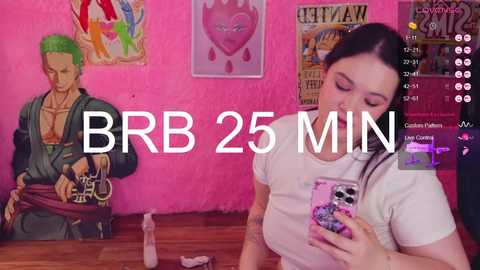 Media: A video of a young woman with long dark hair, wearing a white t-shirt, taking a selfie in a vibrant, pink-themed room adorned with anime posters and a green-haired mannequin.