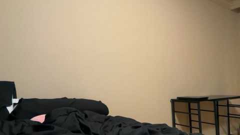 A video of a messy bedroom with a beige wall, black bed covers, a black pillow, a pink sheet, and a wooden nightstand with a black table lamp.