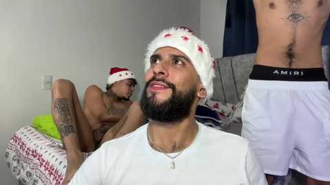 Video of two shirtless men in a bedroom, one with a Santa hat and a necklace, the other with tattoos and a tattooed eye, wearing white underwear.