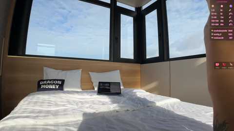 A video of a minimalist bedroom with a large window, showcasing a white bed with a black \"Dragon Hugley\" pillow, a closed laptop, and a vibrant cityscape view.