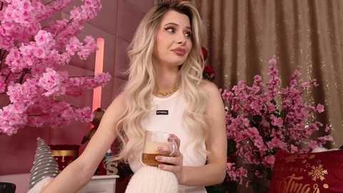 Video of a blonde woman in a white dress, holding a drink, in a room with pink cherry blossom trees and gold curtains.
