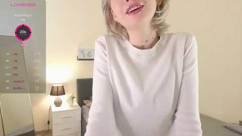 A video of a light-skinned woman with short blonde hair, wearing a white long-sleeve shirt, standing in a minimalist bedroom with a calendar, lamp, and white dresser.