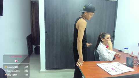 Video of a muscular man in a black tank top and backward cap standing over a seated woman in glasses, white shirt, and red tie, in a dimly lit office.