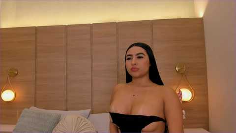 Video of a voluptuous, light-skinned woman with long black hair, wearing a black strapless top that reveals ample cleavage, seated in a modern bedroom with a light wood headboard and wall sconces.