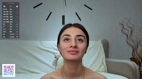 Video of a young woman with dark hair, fair skin, and a slim physique, lying on a white bed in a minimalistic room with a large clock and a decorative vase. QR code in the lower left corner.