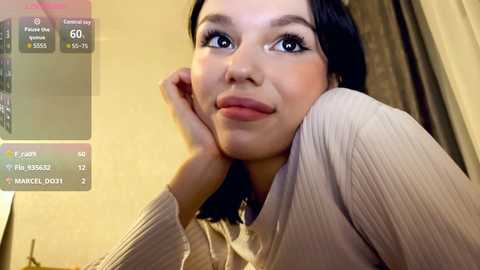 Video of a young woman with fair skin and straight black hair, wearing a beige cardigan, leaning on her elbows, with a casual, neutral expression. Background includes a screen displaying \"Reveal Cam\" and \"Camera Mode\" options.