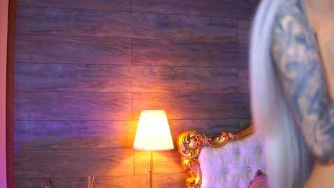 A video of a cozy bedroom corner featuring a wooden wall, a fancy upholstered bed with a blue patterned blanket, and a golden lamp emitting a warm glow.
