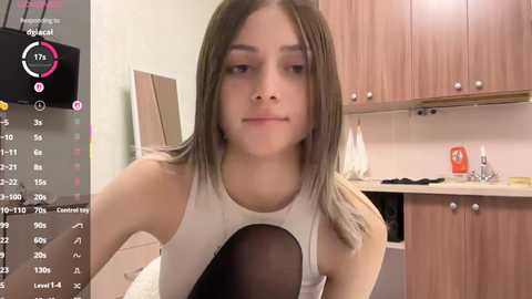 A video of a young woman with straight, medium-length brown hair, wearing a white sleeveless top, sitting in a modern kitchen with light-colored cabinets and a white counter.