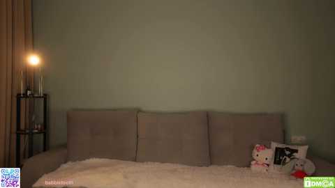 Video of a minimalist bedroom with a beige tufted headboard, a single lightbulb on a black stand, and a plush white teddy bear on a white bedspread.