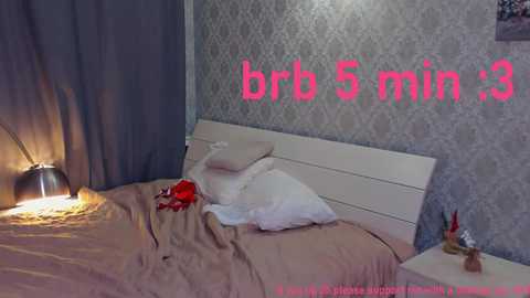 A video of a cozy bedroom featuring a white bed with beige sheets, a red shirt, and a white pillow. Text reads \"brb 5 min 13\" in pink.