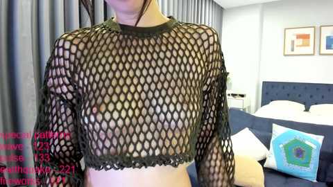 Video of a fair-skinned woman wearing a black fishnet top, revealing her small breasts. Background shows a modern bedroom with a bed, blue pillows, and framed art. Text overlay: \"Rebecca's Instagram Live: Rebecca's Nipples: 21.2k Views.\