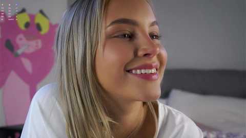 Video of a smiling, light-skinned woman with straight, blonde hair in a white shirt, sitting in a brightly lit bedroom with pink decorations.