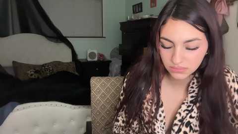 Video of a young woman with long dark hair, wearing a leopard print robe, sitting in a dimly lit bedroom with a black bed, white wall, and a fan on a dresser.