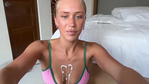 Video of a blonde woman with light skin, wearing a pink sports bra, kneeling on a bed in a bedroom, with semen on her chest.