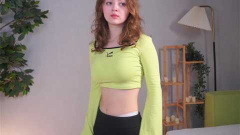 Video of a young, fair-skinned woman with curly red hair, wearing a lime green crop top and black leggings, standing in a modern living room with a green couch, wooden shelves, and a lamp.