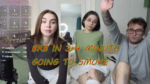 Video of three young people, two women and one man, sitting on a couch in a modern, brightly-lit room. One woman is vaping, the other is smiling, and the man is wearing glasses. Text overlay reads \"BB IN 3-4 MINUTE GOING TO SMOKE.\