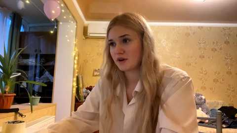 Video of a young woman with long blonde hair, wearing a white blouse, talking in a cozy room with yellow wallpaper, potted plants, and fairy lights.