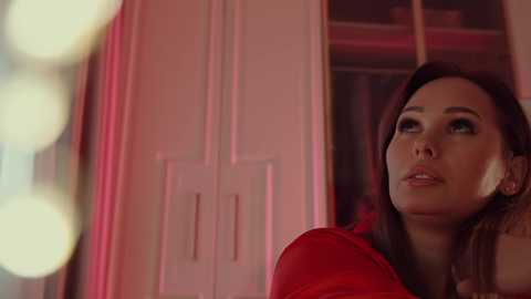 Video of a young woman with light skin and dark hair, wearing a red top, gazing pensively through a window with blurred bokeh lights in the background.