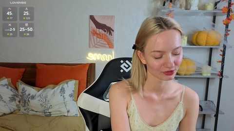 Video of a blonde woman in a yellow tank top, sitting in a black gaming chair with orange pillows, pumpkin decor, and a glowing \"LOVING\" sign in a cozy bedroom.