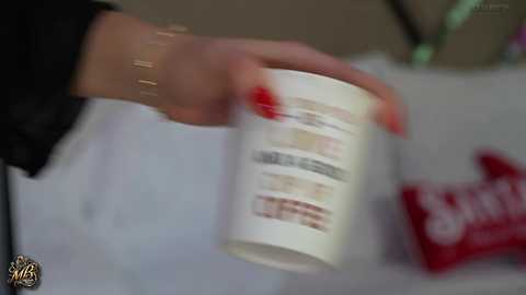 Video of a person's hand holding a white coffee cup with text, red nails, and a black wristband, blurry background with red coffee bag.