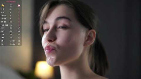 Video of a fair-skinned young woman with brown hair and makeup, pouting and with semen on her lips, indoors with blurred background.