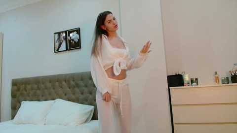 Video of a slim, light-skinned woman with long brown hair in a white satin robe and matching panties, standing in a minimalist bedroom with a white headboard, white bed, and a dresser.
