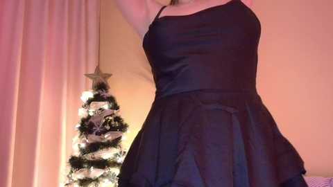 A video of a woman in a black, sleeveless, satin dress with a fitted bodice and flared skirt, standing next to a decorated Christmas tree with white lights and a star topper, in a softly lit room with peach-colored walls.