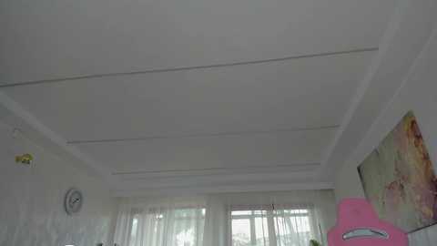 Video of a minimalist, white-walled room with a ceiling fan and white curtains covering a window. A pink toy and a colorful map decorate the right wall.