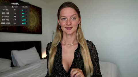 Video of a smiling, light-skinned woman with long blonde hair in a black sheer blouse. She stands in a modern bedroom with a white bed and a large, abstract wall painting.