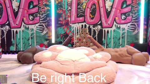 A video of a nude woman reclining on a plush, pink, cloud-shaped pillow, surrounded by more pillows, in a colorful, graffiti-covered room with the words \"LOVE\" in large letters.