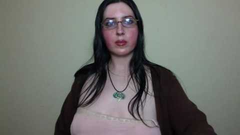 A video of a fair-skinned, long-haired woman with glasses, wearing a beige top and a brown cardigan, against a plain green background. She has a neutral expression.