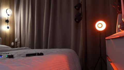 Video of a dimly lit bedroom with beige curtains, a gray quilted bed, a small nightstand with a lamp, a red glowing spotlight, and a black remote control on the bed.