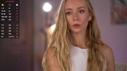 Video of a blonde woman with long, wavy hair, wearing a white sleeveless top, standing indoors against a blurred background.