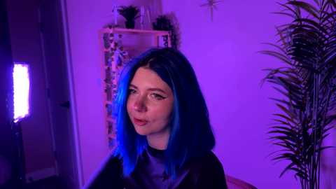 Video of a young woman with blue hair, wearing a black latex dress, standing in a dimly lit room with purple lighting, potted plants, and a shelving unit in the background.