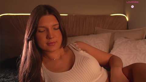 Video of a serene young woman with long, straight brown hair, wearing a white ribbed tank top, lying on a bed with beige pillows and a gray blanket.