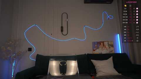 Video of a dimly lit living room with a dark green sofa, a white vase, and a blue neon heart-shaped light fixture on a white wall.