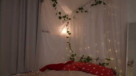 Video of a cozy, dimly lit bedroom with white sheer curtains, fairy lights, and green ivy hanging from the ceiling, creating a whimsical atmosphere. A red pillow with white butterflies is visible on a bed, and soft, warm lighting enhances the romantic ambiance.