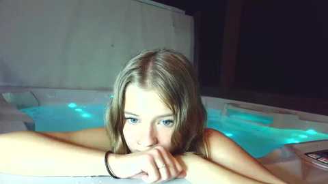 Video of a young Caucasian woman with light brown hair, blue eyes, and fair skin, lying in a lit indoor hot tub, looking contemplative, with dimly lit background.