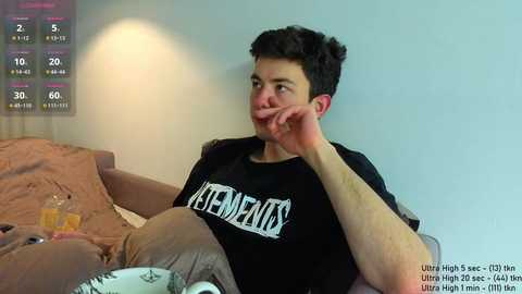 Video of a young man with short dark hair, wearing a black \"PENISHEAD\" shirt, sitting on a couch with a beige blanket, looking at his phone.