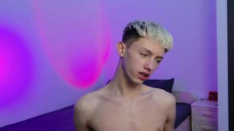 Video of a shirtless, slender young man with bleached blonde hair and a pale complexion, looking slightly downcast. Background features a bed with dark sheets, purple and pink lighting, and a nightstand.