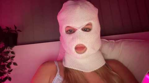 Video of a light-skinned woman with long blonde hair wearing a white balaclava and a light blue lace top. She's seated on a white couch with pink lighting.