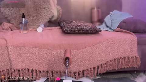 A video shows a messy bedroom with a bed covered in a pink shaggy blanket, featuring a large, realistic pink dildo plugged into an electrical socket, surrounded by scattered items including a bottle of lotion, a phone, and a denim jacket.
