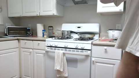 A video of a small, tidy kitchen featuring white cabinets, a microwave, a white stove with a white towel hanging, and a person in a white shirt partially visible on the right.