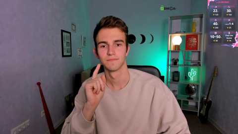 A video of a young man with short brown hair in a beige sweater, making a \"peace\" sign, in a cozy room with a green light, shelves, guitar, and a TV screen displaying game stats.