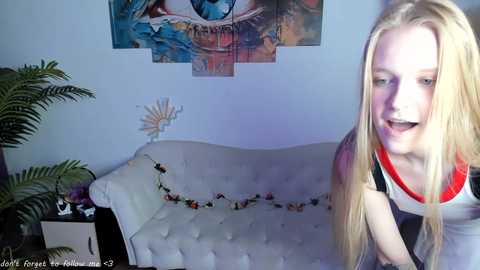 Video of a young blonde woman with long hair, wearing a red and white top, standing near a white tufted couch adorned with a garland, in a colorful, abstract art-filled room.