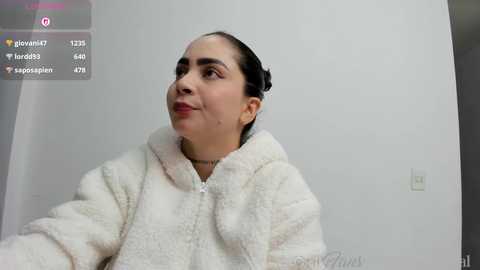 Video of a young Latina woman with dark hair in a bun, wearing a white fluffy robe, standing against a plain white wall, with social media icons in the top left.